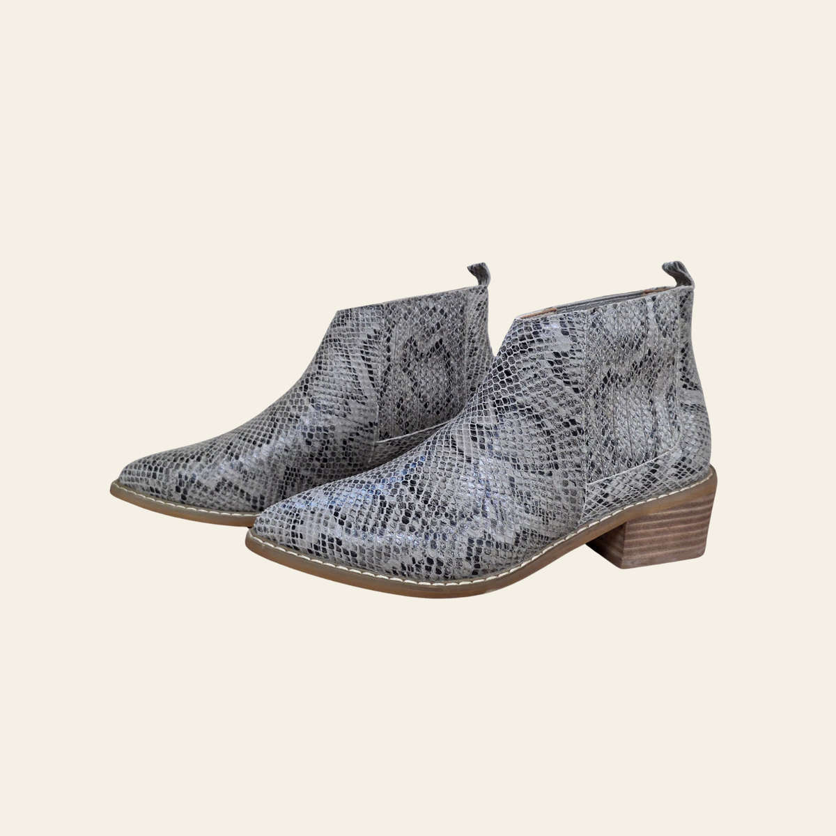 A pair of women's ankle boots featuring a gray and black snakeskin pattern, with a low brown block heel and white contrast stitching, shown against a solid cream background.