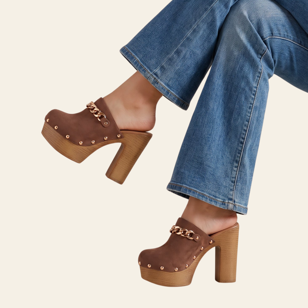 A person wearing brown platform clog-style mule heels with a chunky block heel, studded trim, and a gold-tone chain detail across the top, paired with blue jeans on a light cream background.