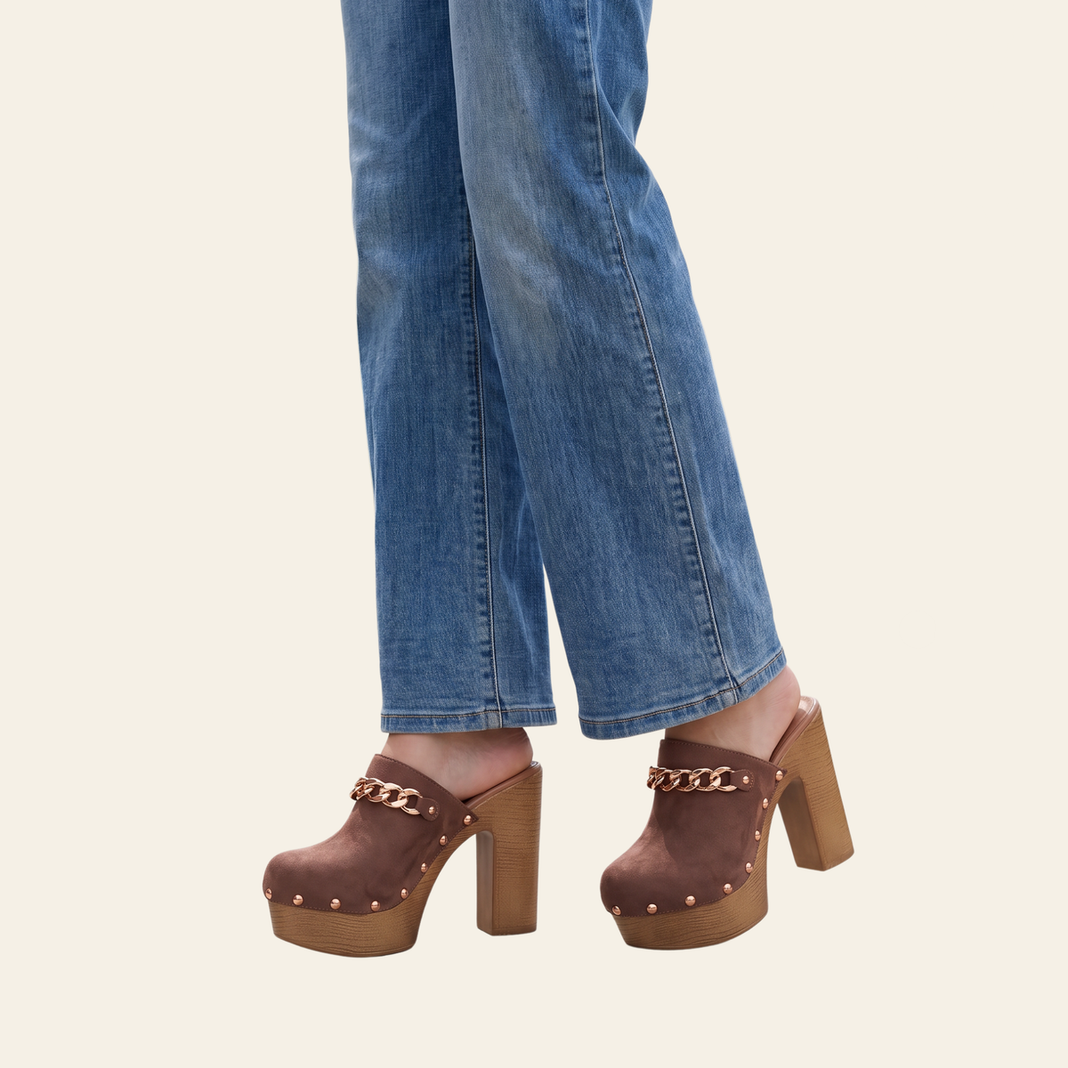 A person wearing brown suede clog-style platform sandals with gold-tone chain straps and studs, paired with blue jeans, shown from the calves down against a plain light beige background.