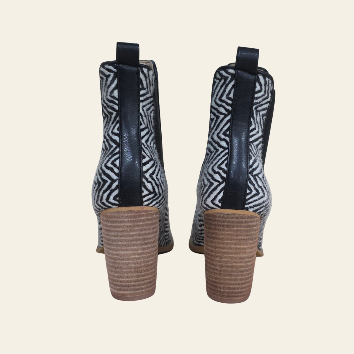 A pair of women's ankle boots shown from the back, featuring a black and white geometric patterned fabric, black leather side panels, and chunky brown stacked block heels, set against a solid light beige background.
