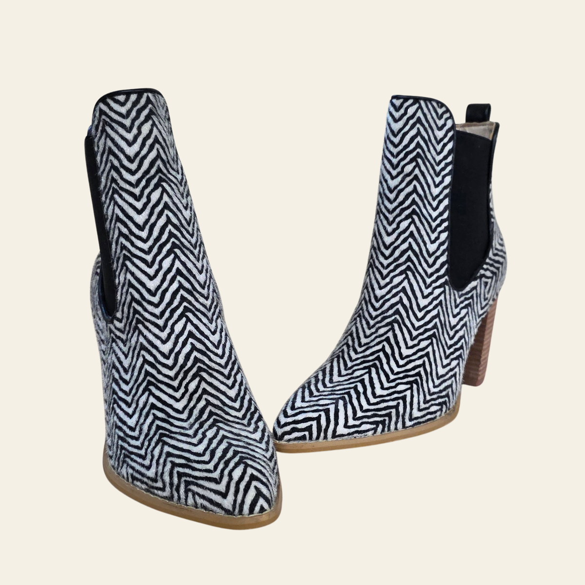 A pair of women's ankle boots featuring a black and white herringbone pattern on the front, with black elastic side panels and brown block heels, set against a solid light beige background.