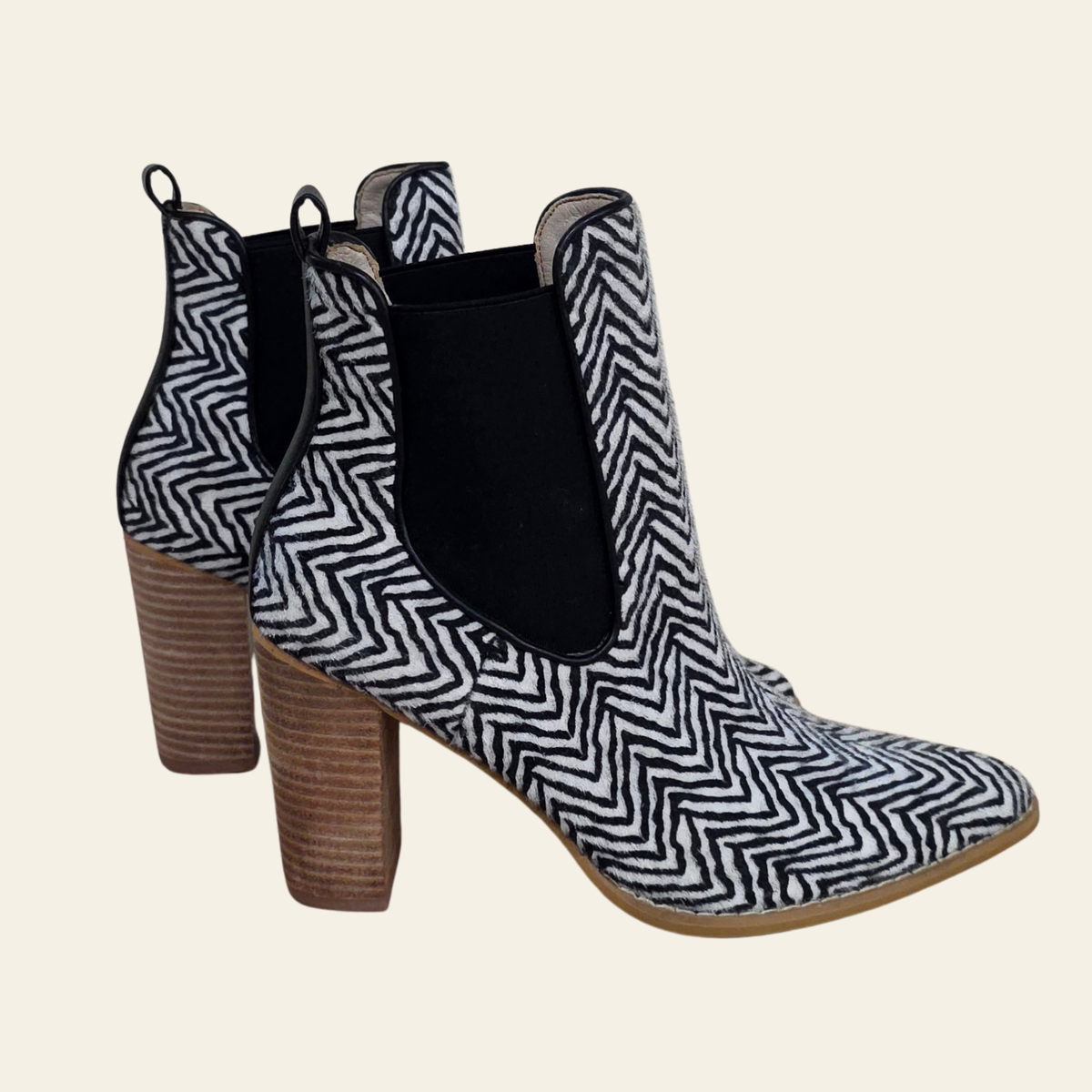 A pair of women's ankle boots featuring a black and white herringbone pattern on the toe and heel, with black elastic side panels and a brown block heel, shown against a solid light beige background.
