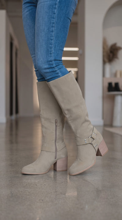 Model wearing taupe knee-high harness boots with silver stud accents and block heels, moving to show the boots from different angles.