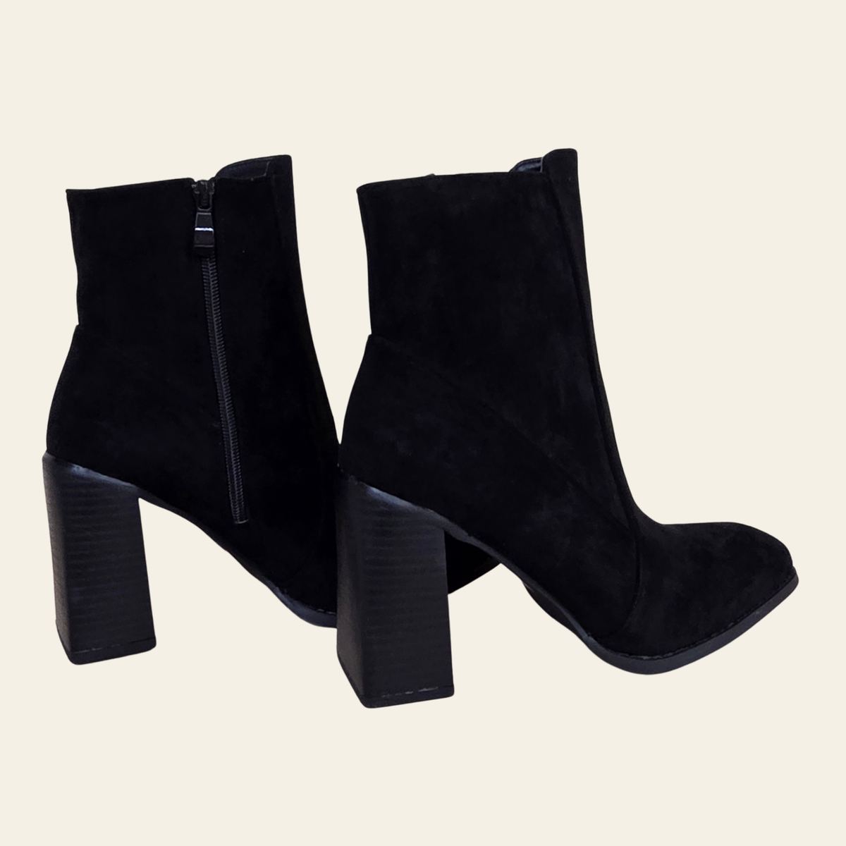 A pair of black suede ankle boots with block heels and side zippers, shown from the back and side against a light cream background.