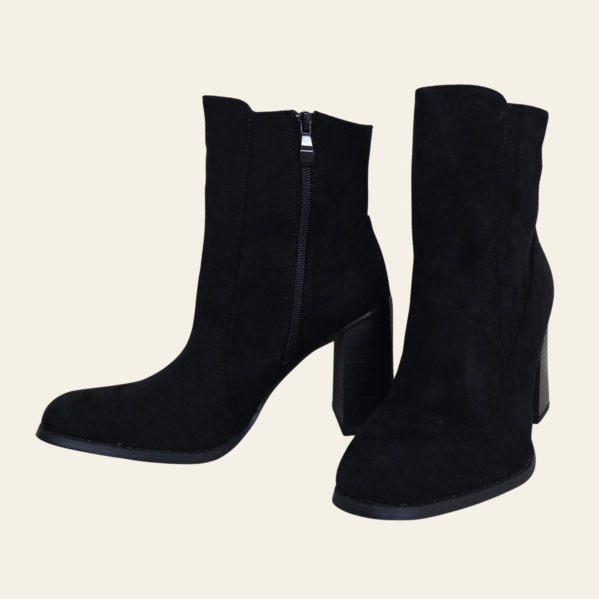 A pair of black suede ankle boots with block heels and side zippers, displayed against a solid light beige background.