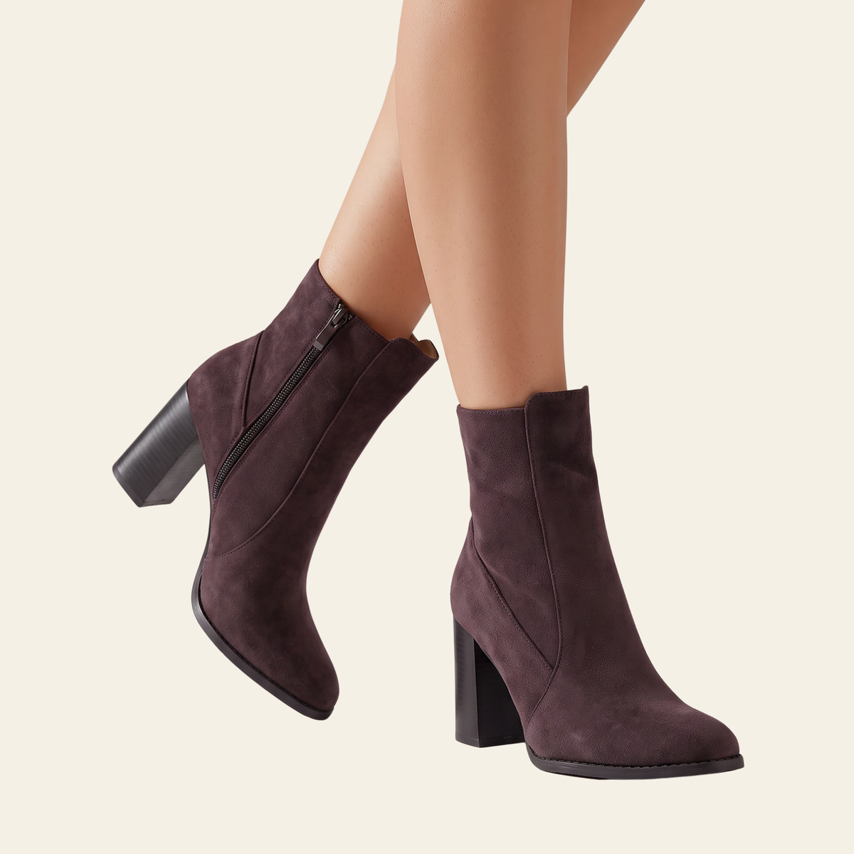 A pair of women's ankle boots in brown faux suede, shown on a model's feet against a solid light beige background. The boots feature a chunky block heel, a side zipper, and a pointed toe.