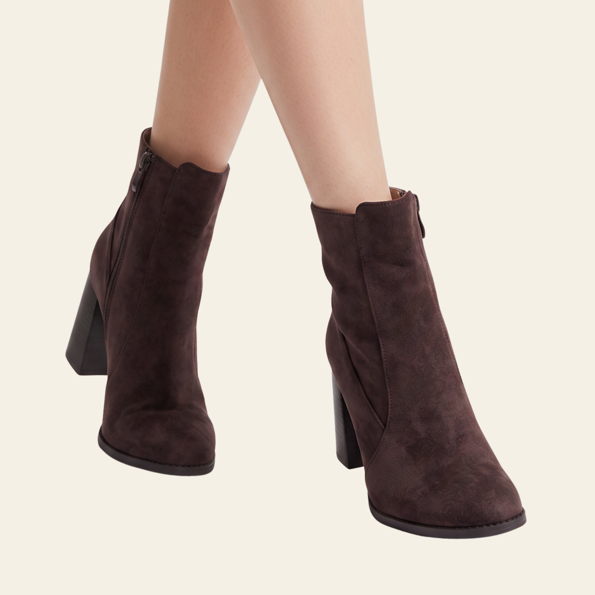 A pair of dark brown suede ankle boots with block heels, shown on a model's feet against a light cream background.